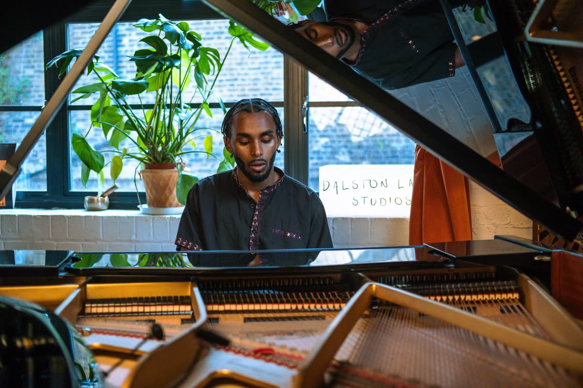 Judah at a grand piano with plants in the window behind, seen past the open lid