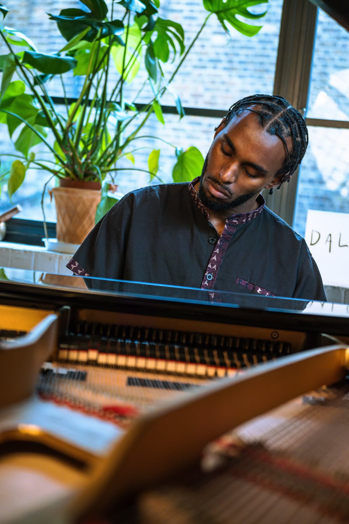 Judah playing a grand piano, plants in the window behind