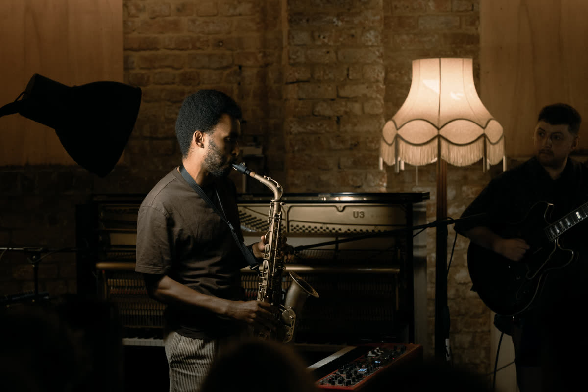Judah playing saxophone under a fringed lamp, a guitarist beside him